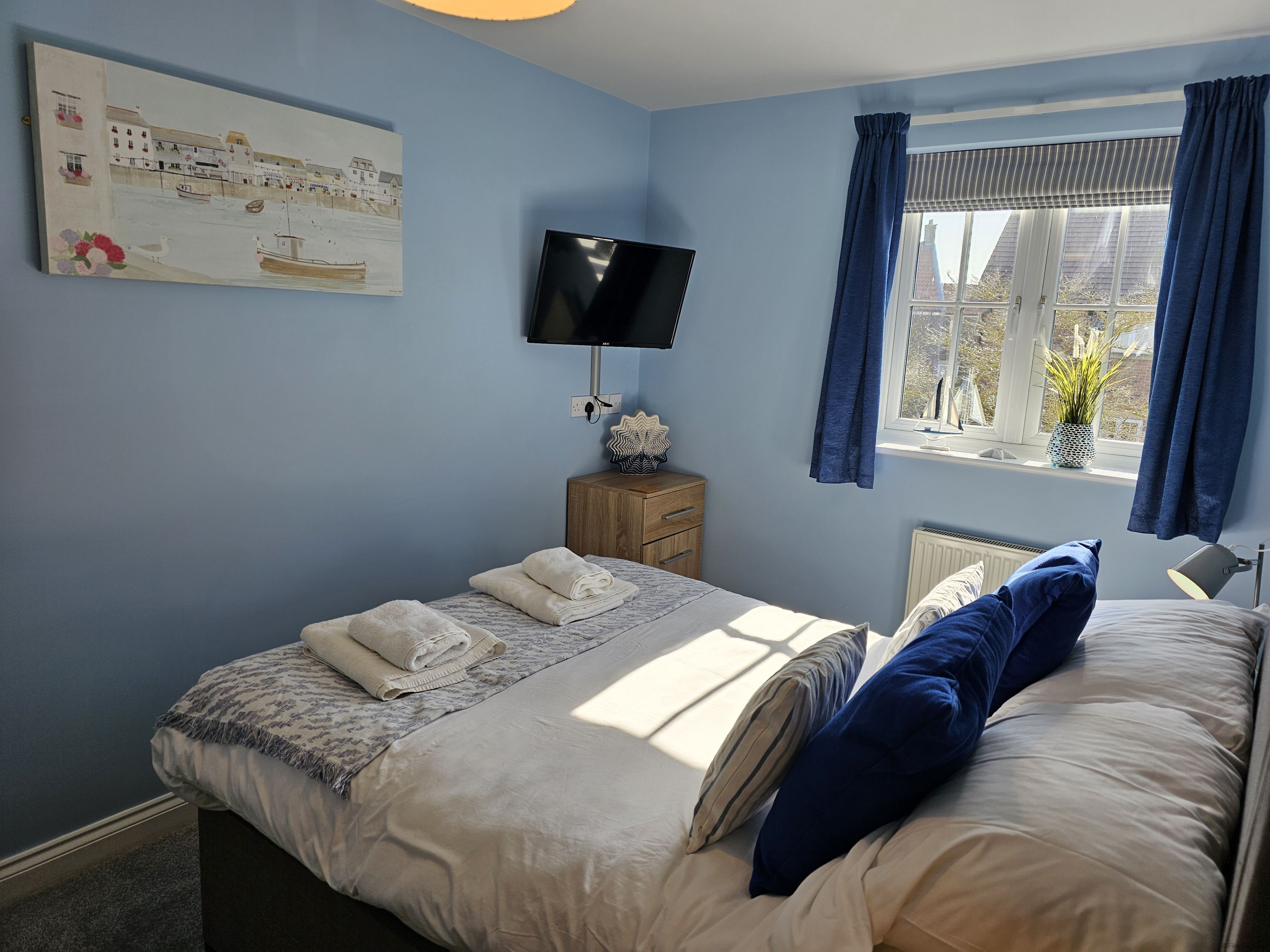 Een slaapkamer met een bed, een flatscreen televisie, een raam met gordijnen, een nachtkastje en een aan de muur hangend kunstwerk.