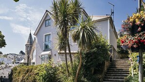 Une maison à deux étages dotée d’un balcon, entourée d’une végétation luxuriante et surplombant un ciel d’un bleu éclatant.