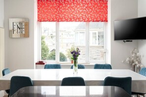Un comedor con una mesa larga, sillas, un reloj, una ventana con una persiana floral roja y un televisor montado en la pared.