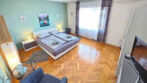 Ein Schlafzimmer mit Bett, Nachttischen, einer Lampe, einem Sessel, einem Fernseher und einem Fenster mit Vorhängen.