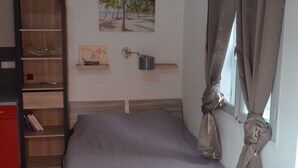 Een slaapkamer met een bed, een houten plank met boeken, een wandplank met een kaart en een pot, en een raam met gordijnen.