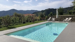 Una piscina rectangular con agua azul cristalina, rodeada por un muro de piedra y una terraza con baldosas. Hay cuatro sillas de descanso y una pequeña mesa cerca de la piscina. El fondo muestra un paisaje montañoso con árboles y algunos edificios.