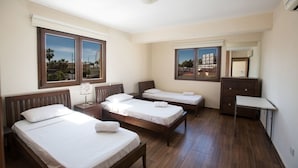 Habitación de hotel con tres camas individuales, un aparador, un espejo y dos ventanas con vistas a palmeras y edificios.