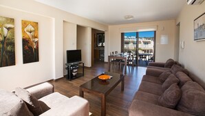 Una sala de estar con un sofá, una mesa de centro y un televisor. Cuenta con un comedor con una mesa y sillas. La habitación tiene vista a edificios.