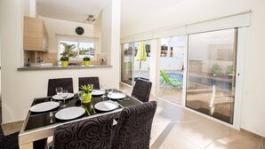 Zona de menjador amb taula preparada per a quatre, cadires amb disseny estampat, cuina amb armaris de fusta i porta corredissa de vidre que dona a una zona exterior amb piscina i gandules.