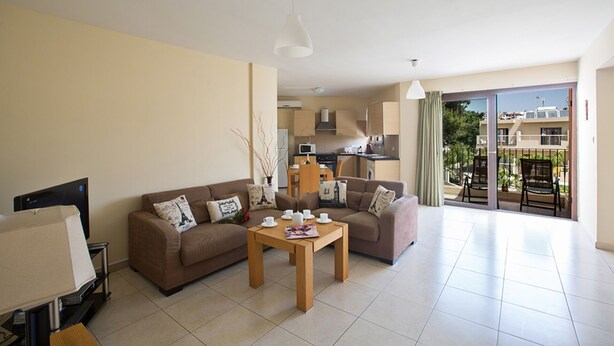 A living room with a sofa, coffee table, and a TV. There is a kitchen area with cabinets and a dining table. The room has a balcony with a view of buildings and greenery.