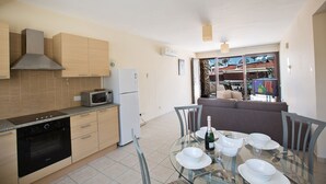 A compact kitchen with a microwave, oven, and refrigerator. A dining area with a glass table and chairs is visible, with a sofa and a TV in the background. - Picture This (Ayia Napa)