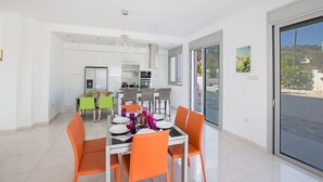A modern dining area with a glass table and orange chairs, a kitchen in the background, and a sliding glass door leading to a balcony. - Luxury 5 Bedroom Villa with Private Pool, Protaras Villa 1230 (Protaras)
