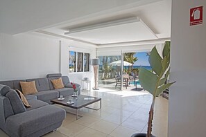 Un salon moderne avec un canapé gris, une table basse et une grande fenêtre donnant sur une piscine et la mer.