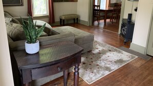 A living room with a sofa, ottoman, and a coffee table. There is a painting on the wall, a plant, and a dining area in the background. - Summer And Track Season Rental (Saratoga Springs)