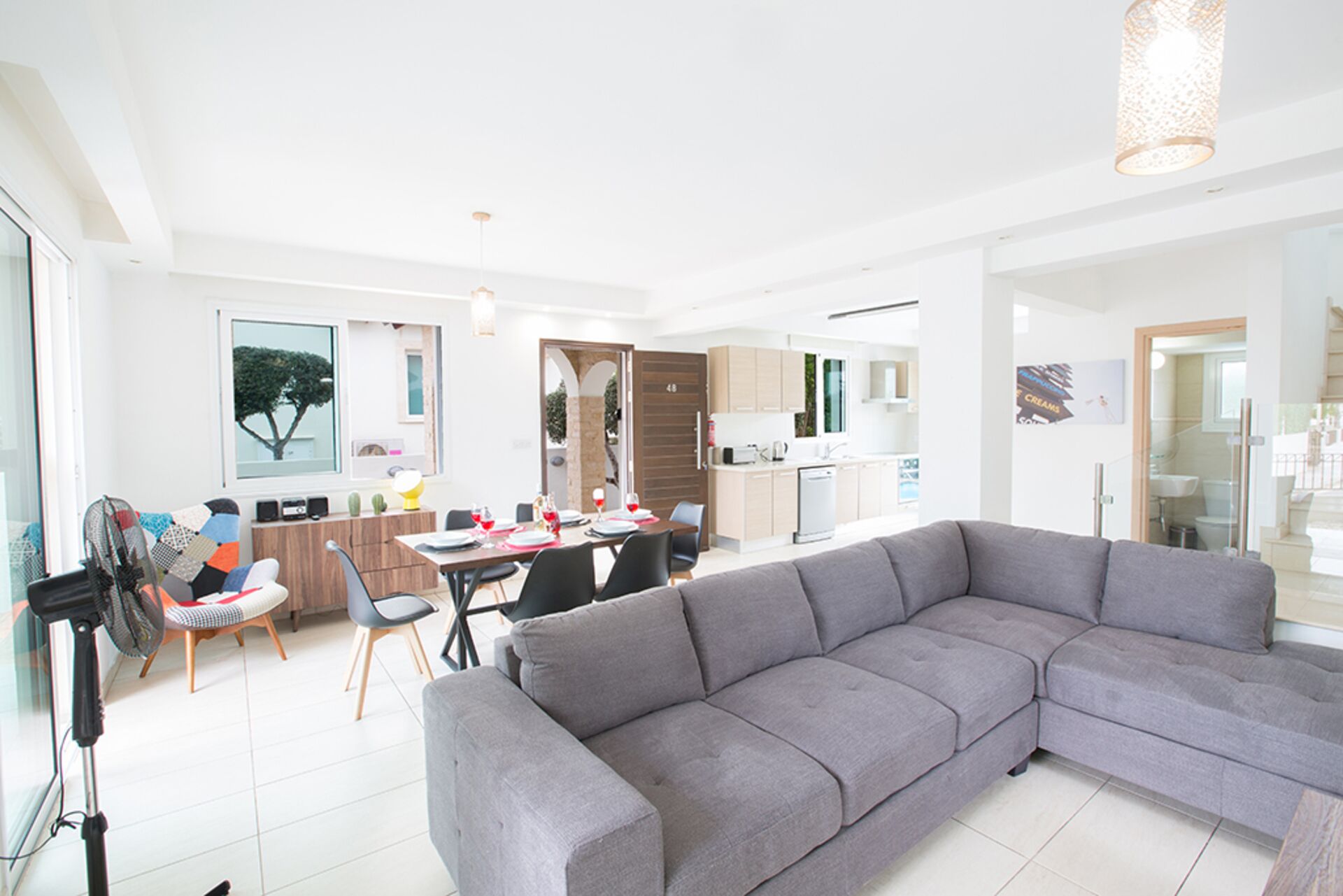 A modern living room with a grey sectional sofa, a dining area with a table and chairs, and a kitchen in the background.