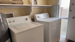 Une buanderie équipée de deux machines à laver et deux sèche-linge blancs, d’un fer à repasser gris et d’une porte donnant sur une autre pièce.