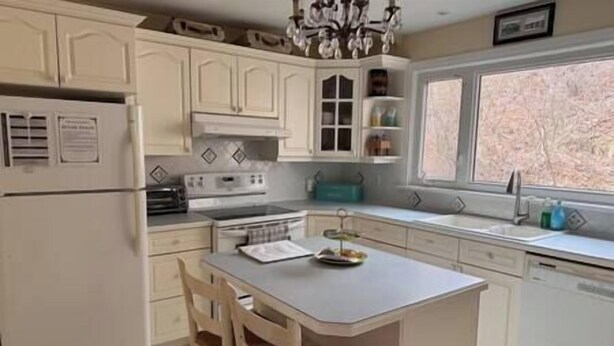 Une cuisine avec des meubles blancs, un îlot central, un réfrigérateur, une cuisinière et une fenêtre donnant sur des arbres.