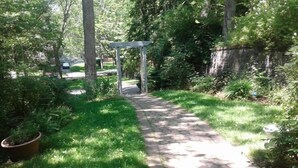 Un chemin de jardin pavé qui mène à une pergola, elle-même entourée d’une végétation luxuriante et d’arbres.