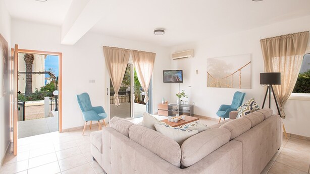 A spacious living room with a beige sofa, two blue armchairs, a coffee table, and a TV mounted on the wall. - How to Rent Your Own Private Luxury Holiday Villa in Protaras for Less than Basic Hotel (Protaras)