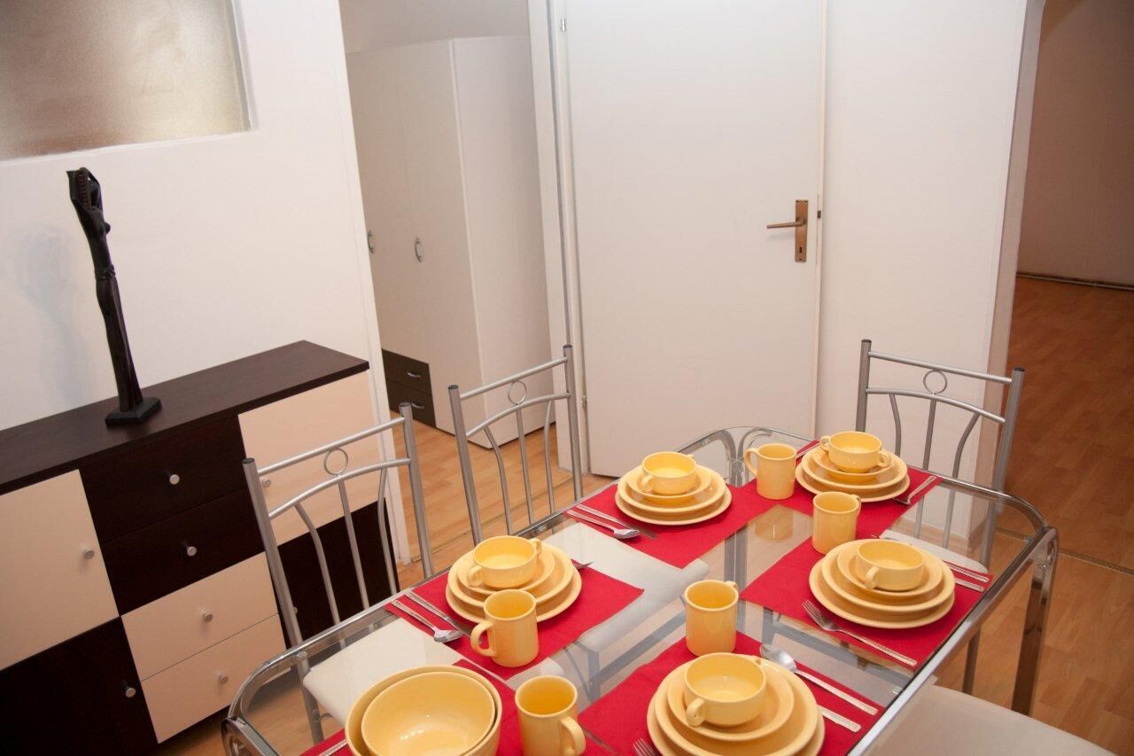 A dining area with a glass table set for four, surrounded by metal chairs. A dark-colored dresser is to the left, and a white door is in the background.