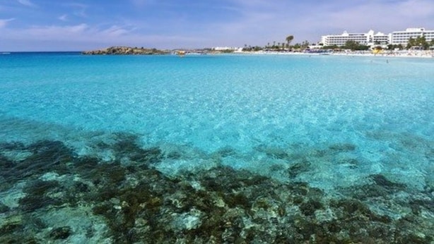 Klares blaues Meer mit steinigem Untergrund, ein Sandstrand und ein Hotel im Hintergrund.