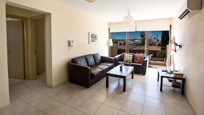 Ein Wohnzimmer mit einer Couch, einem Couchtisch und einem kleinen Tisch mit einer Lampe. Links befindet sich eine Tür und ein Balkon mit Blick auf die Stadt.