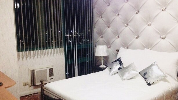 A hotel room with a bed, two bedside lamps, a window with blinds, and a view of the city at night. - 1-BR Robinson Place Residences, 68 sqm. Condo (Ermita Manila)