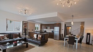 Una sala de estar moderna con un sofá, una mesa de centro y un comedor con sillas. Se observa una cocina con refrigerador y microondas.