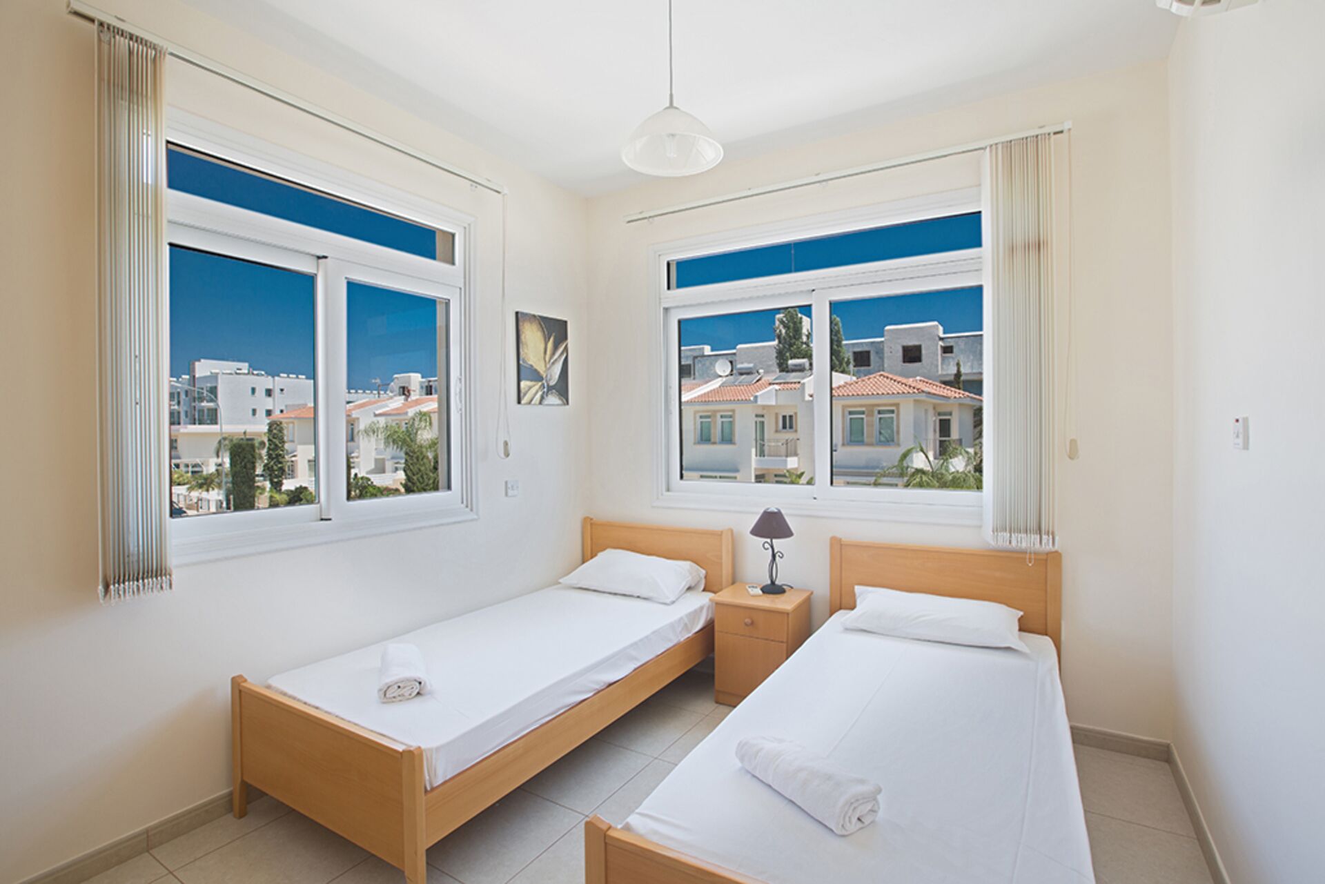 Habitación de hotel con dos camas individuales, una mesita de noche pequeña con lámpara y ventanas amplias que ofrecen vistas de la ciudad.
