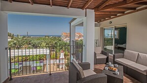 A balcony with wicker furniture, a table with a plate of food, and a view of the sea and greenery.