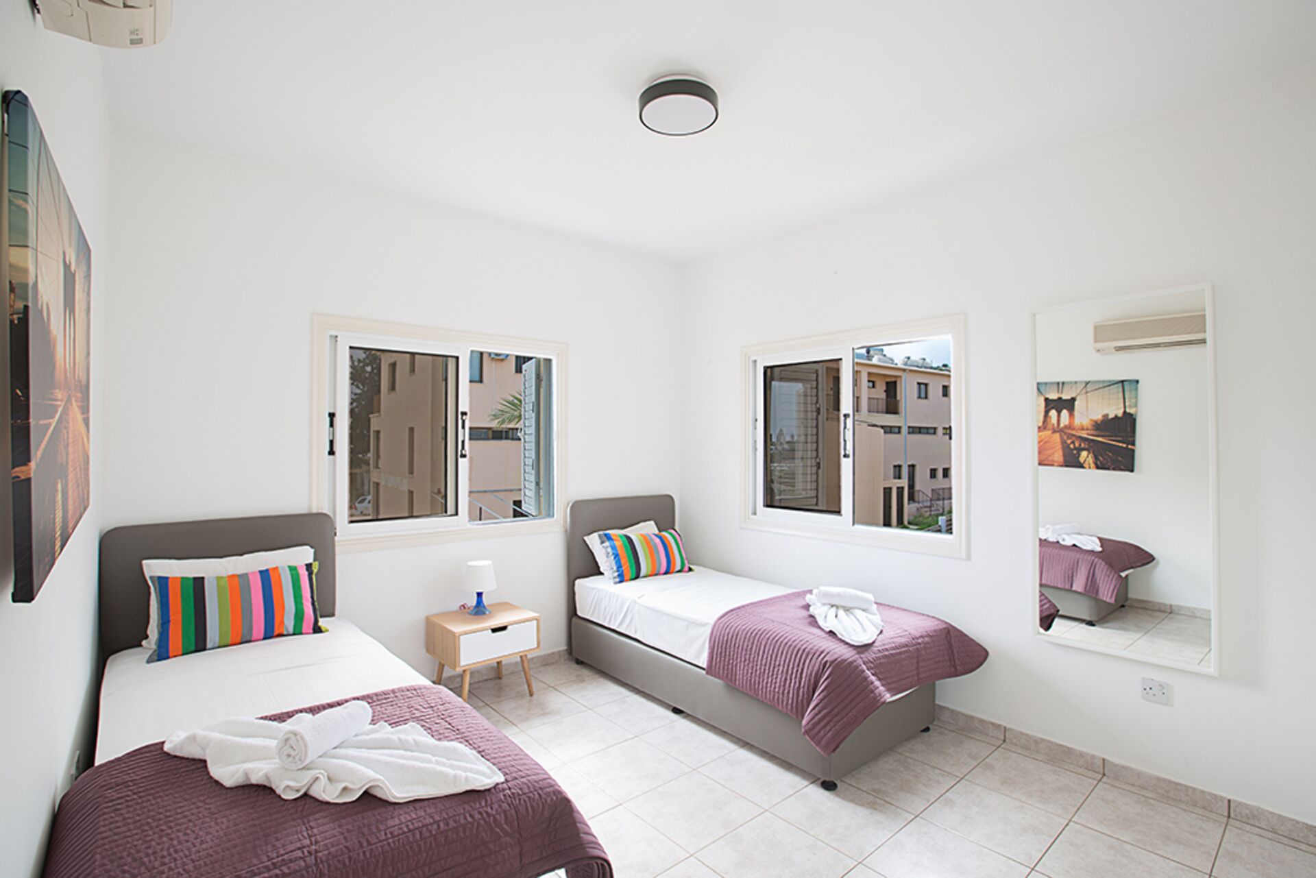 A modern bedroom with two beds, a small bedside table, a mirror, and large windows offering views of buildings and a bridge.