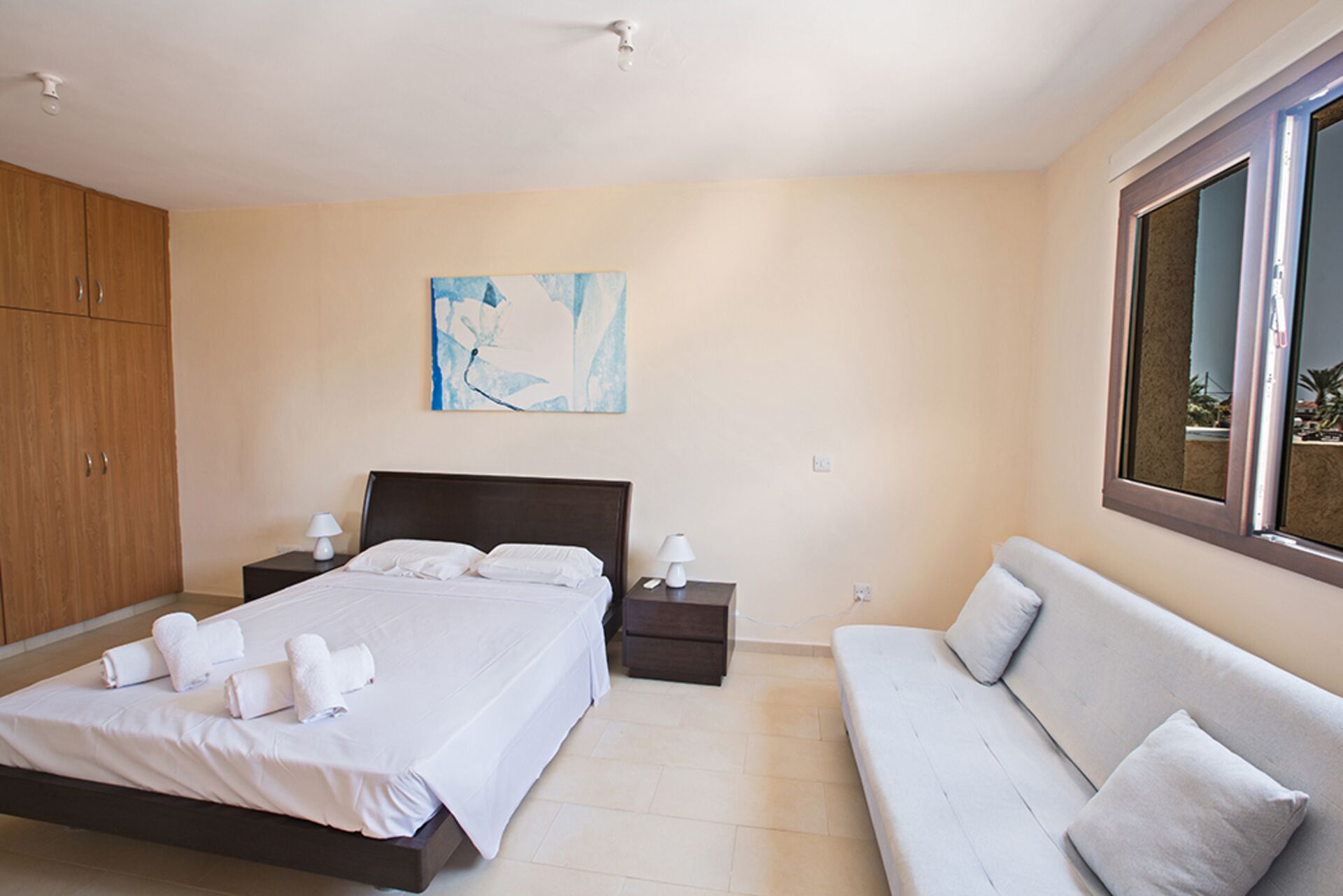 Quarto com uma cama grande, mesa de cabeceira com abajur, sofá e janela.
