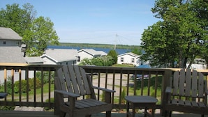 Une terrasse en bois avec deux fauteuils Adirondack et une petite table, donnant sur un lac avec des maisons en arrière-plan.
