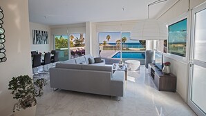 Un salon moderne avec un canapé gris, un coin repas avec des chaises, une grande fenêtre donnant sur une piscine et des palmiers, et un luminaire au plafond.