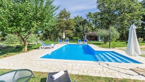 Une piscine rectangulaire à l’eau bleue, entourée d’une terrasse en pierre. On y trouve deux chaises longues bleues et un parasol blanc.