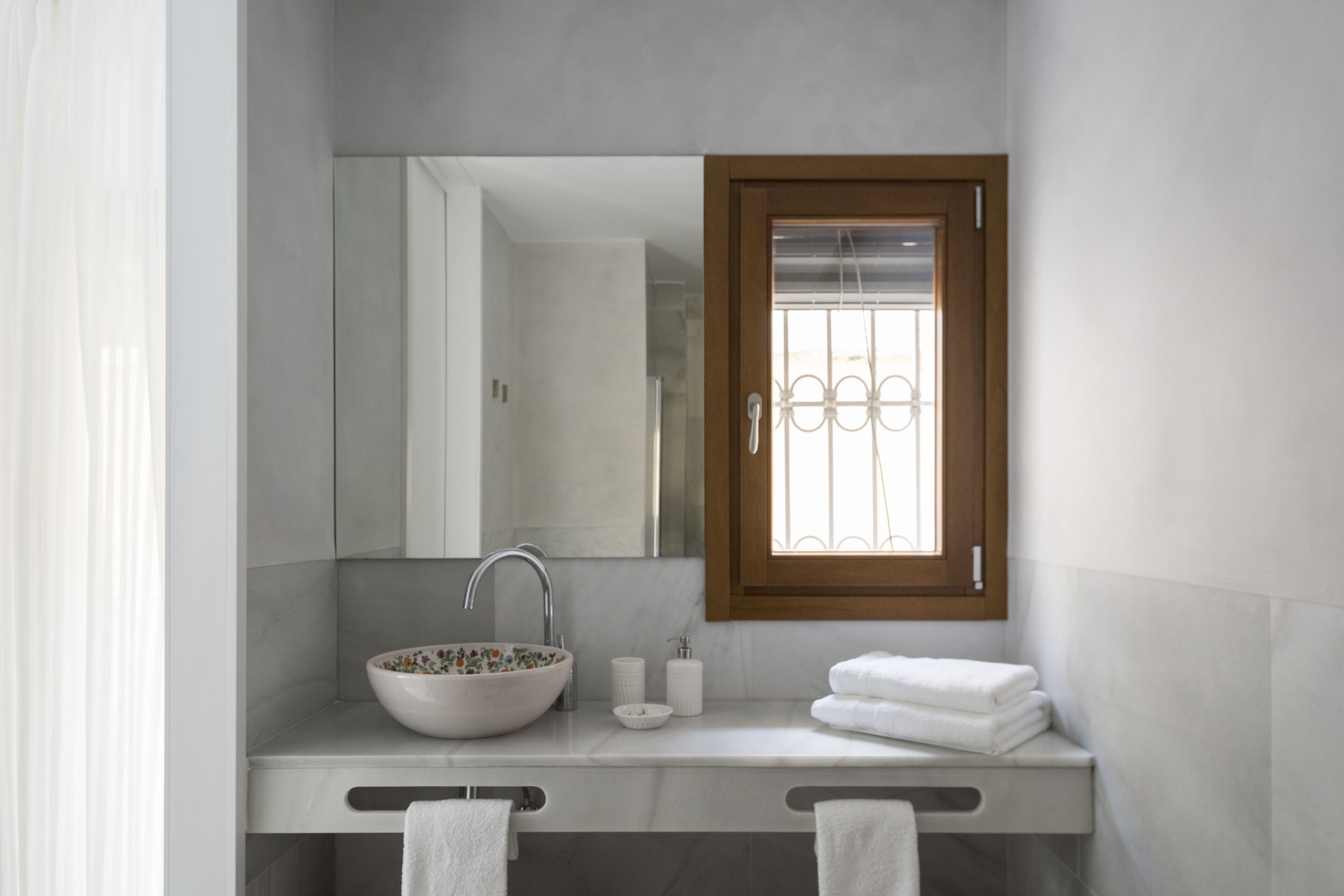 Une salle de bain moderne avec un grand miroir, un lavabo sur colonne et une fenêtre avec des barreaux.
