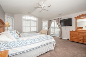 A bedroom with a bed, a ceiling fan, a TV, a dresser, and large windows.