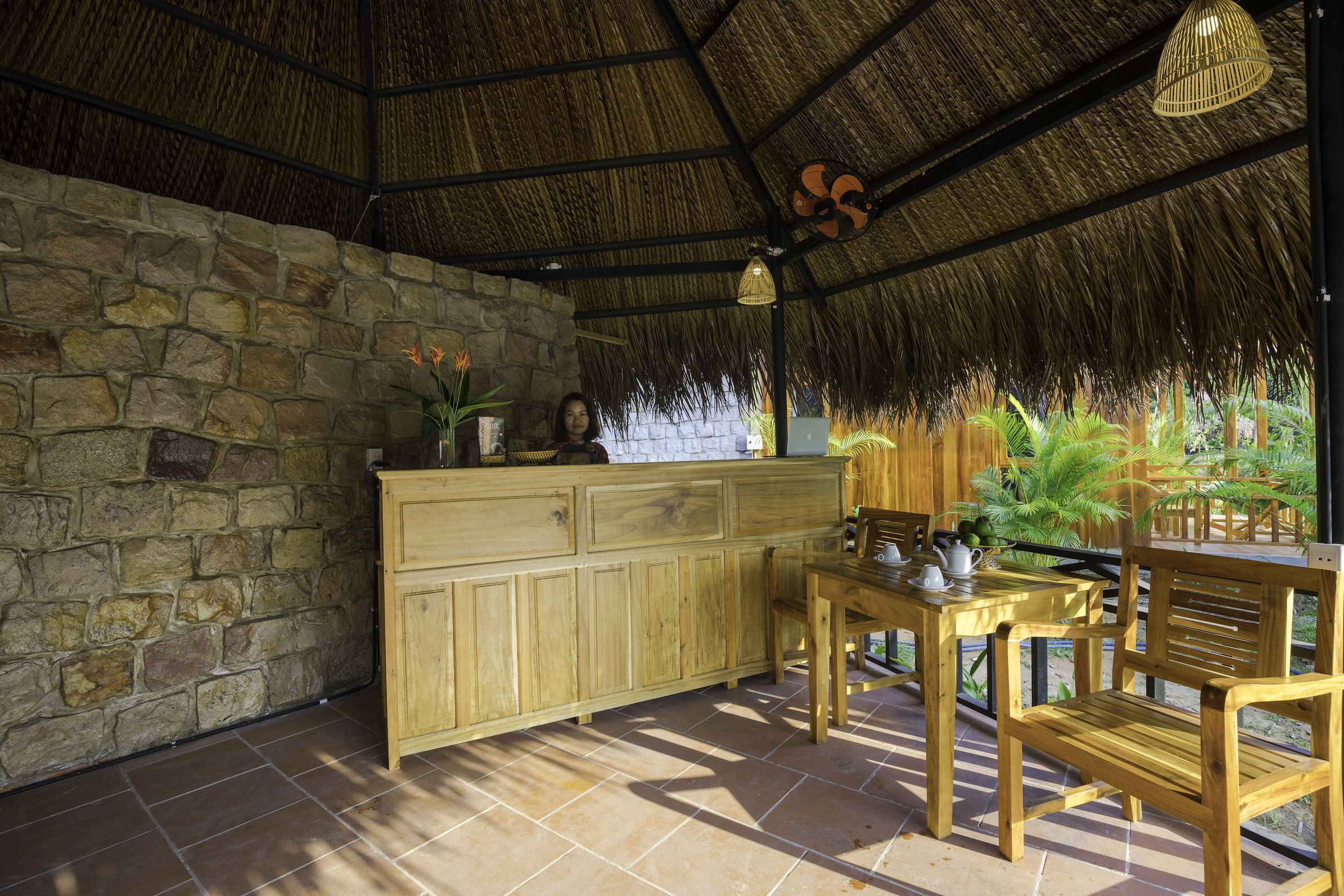 A rustic wooden bar with a stone wall backdrop, a thatched roof, and a view of lush greenery outside.