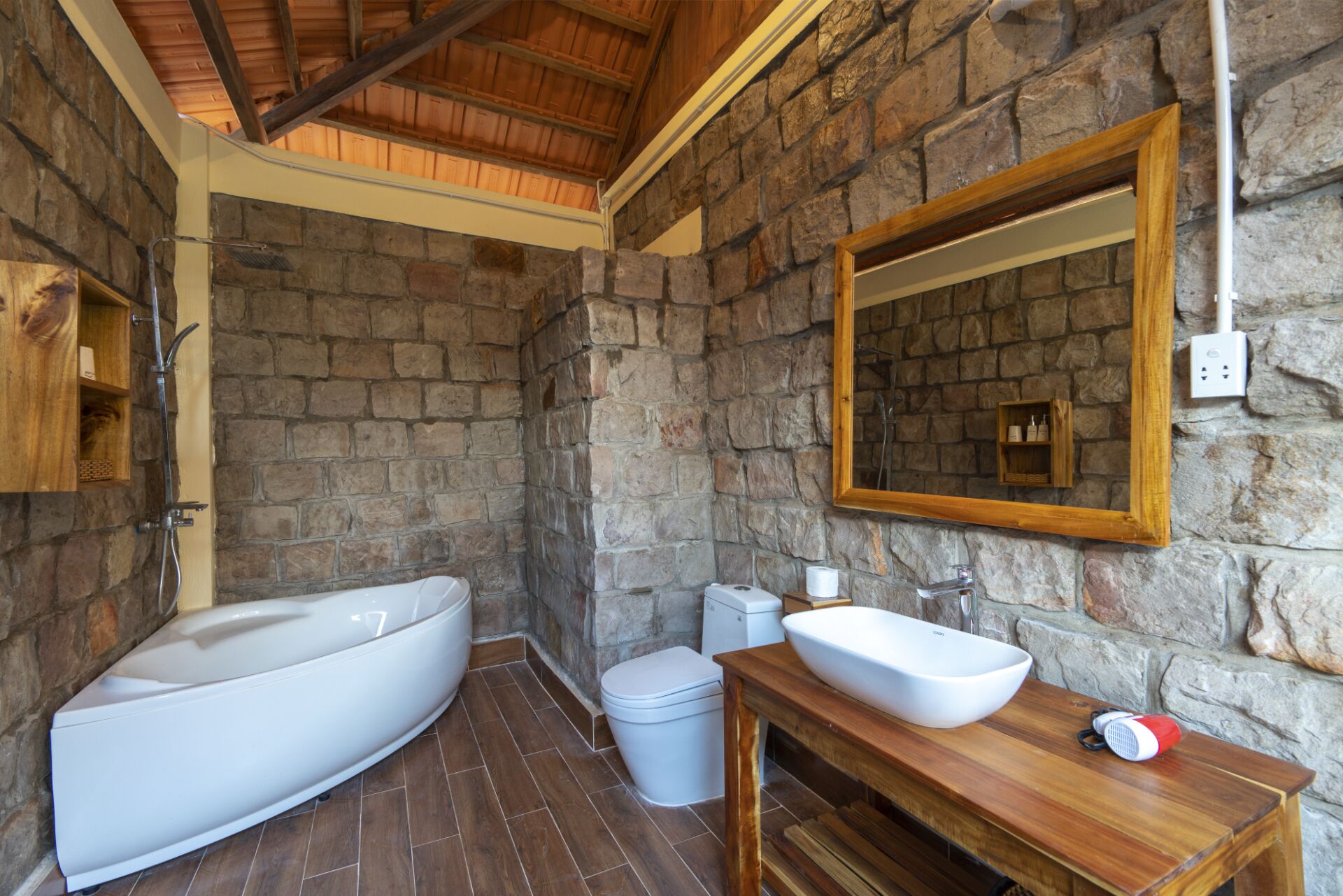 A bathroom with a stone wall, a wooden vanity, a large bathtub, and a toilet.