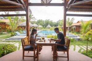 Ein Holztisch mit zwei Stühlen, ein Pool und ein Garten mit Palmen und strohgedeckten Gebäuden.