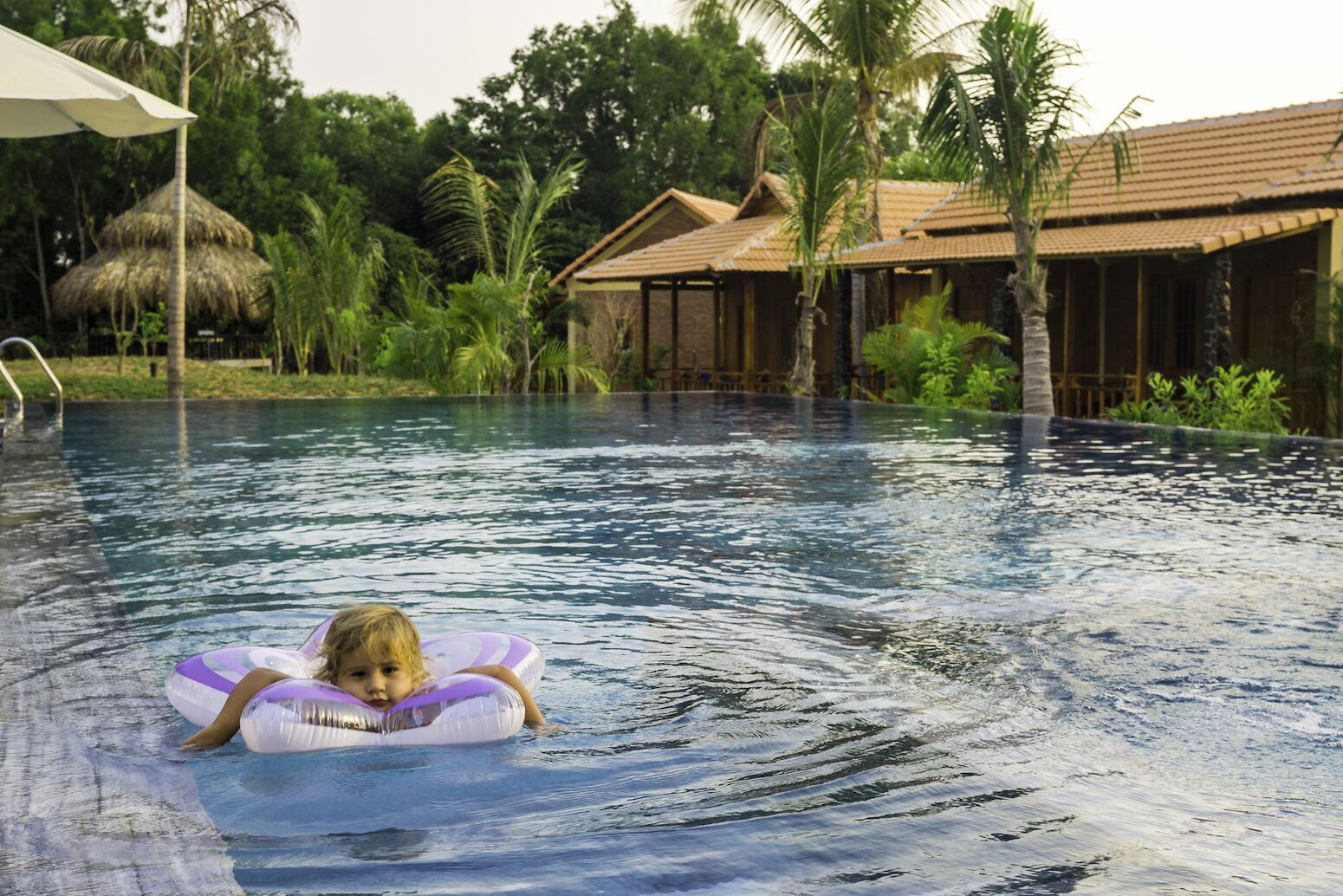 A pool area with a child on a float, surrounded by tropical vegetation and a thatched-roof structure.
