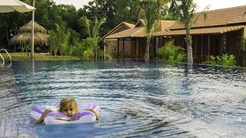 Uma área de piscina com uma criança em um boião, cercada por vegetação tropical e uma estrutura com telhado de palha.