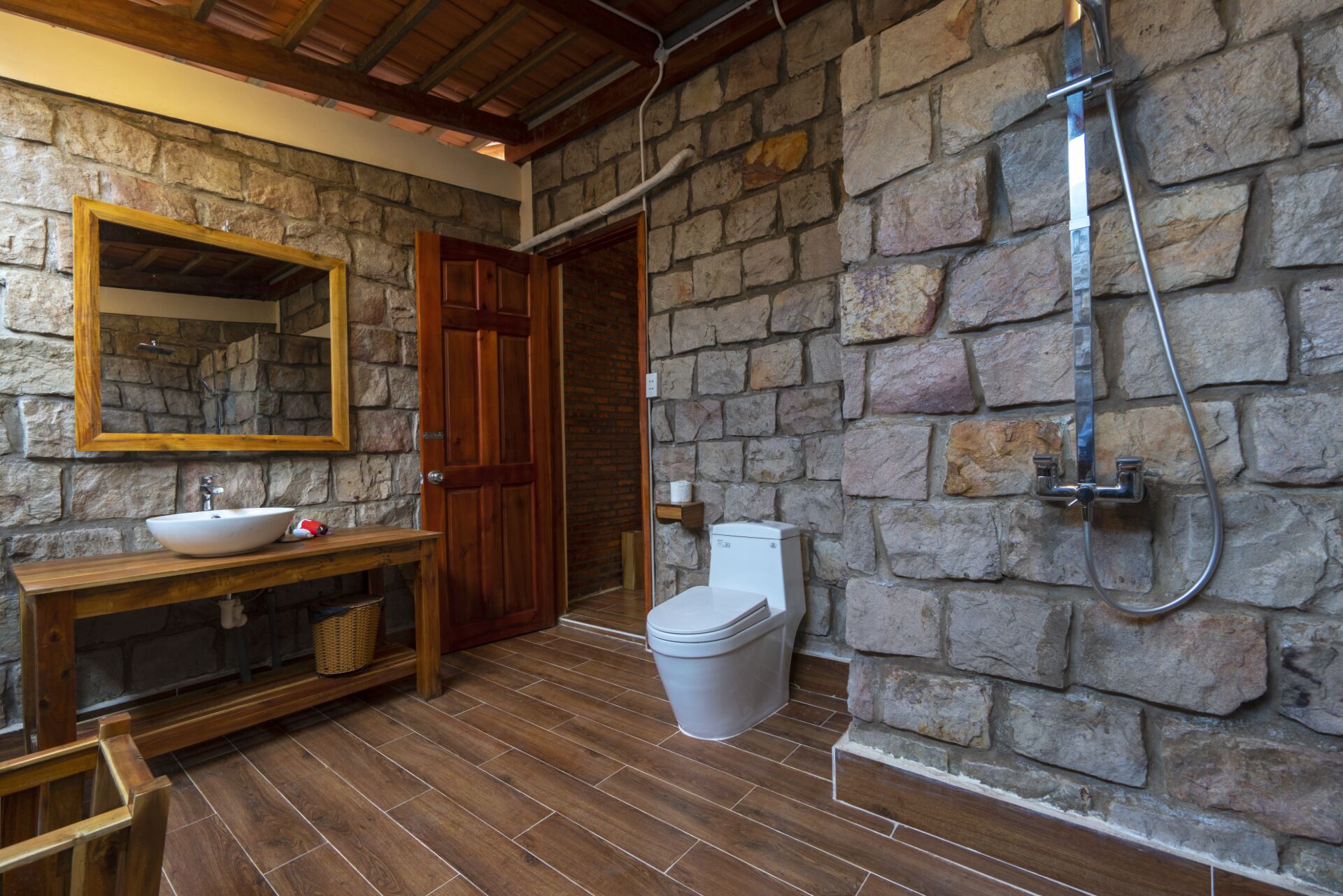A bathroom with a stone wall, a wooden vanity, a sink, a toilet, and a shower.