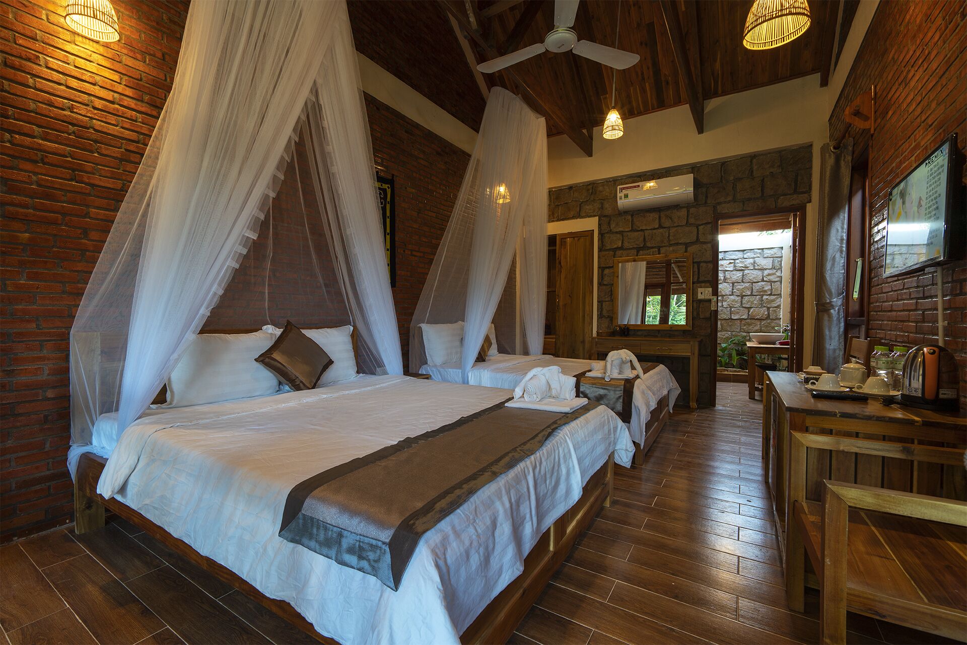 A room with a large bed, two smaller beds, a wooden desk, and a stone wall.