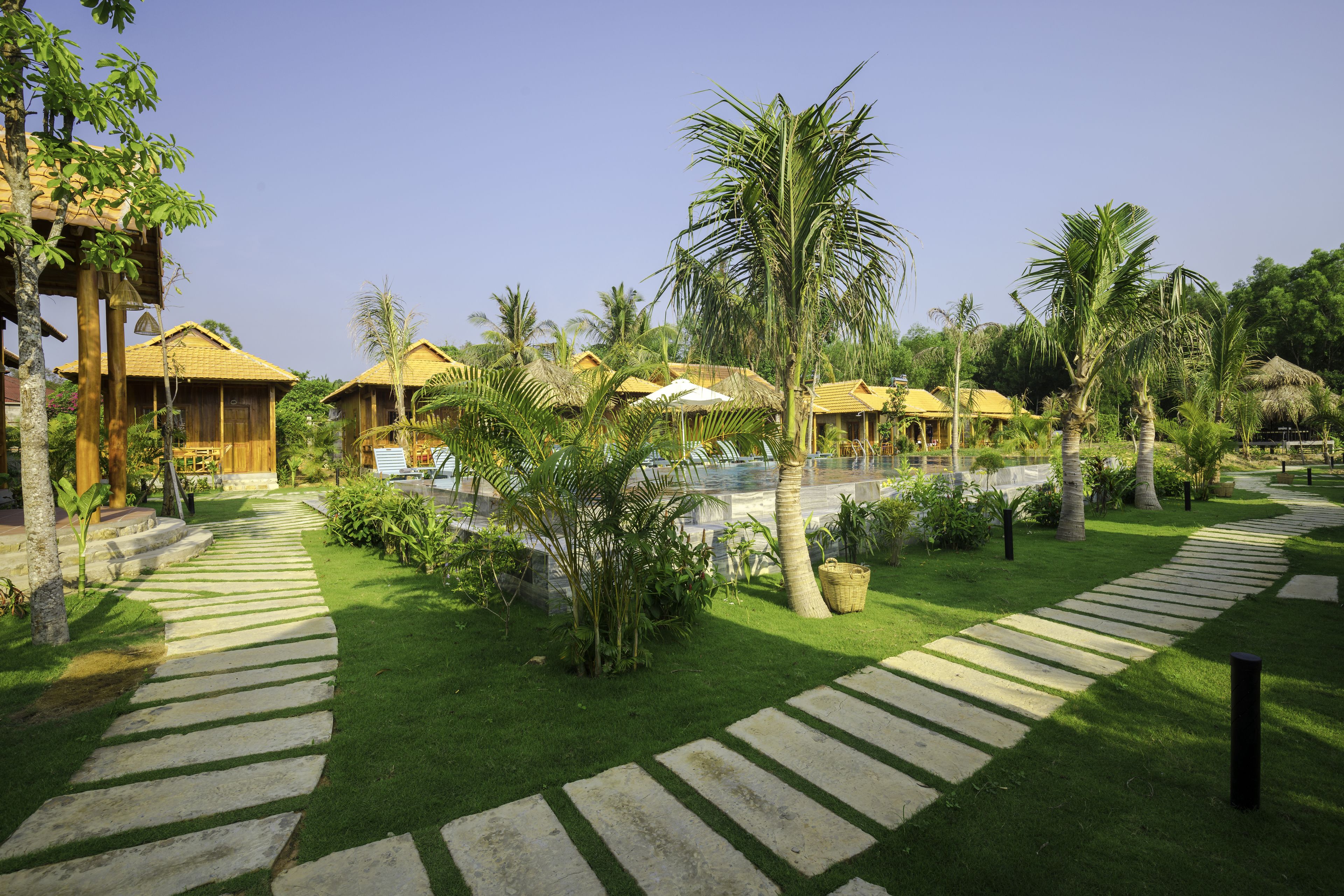 A resort with thatched-roof cottages, a central pool, and a well-maintained garden with palm trees and a stone pathway.