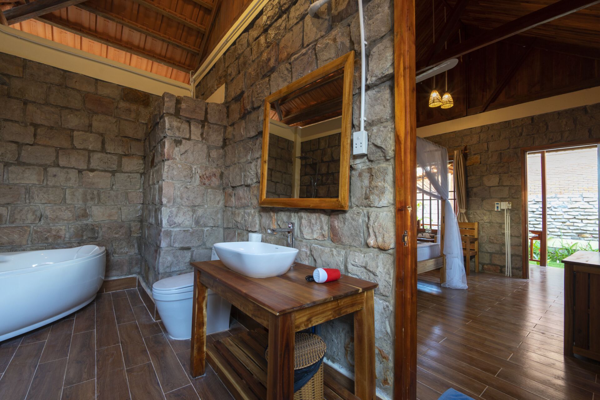 Un baño rústico con pared de piedra, tocador de madera, lavabo blanco y un espejo grande.
