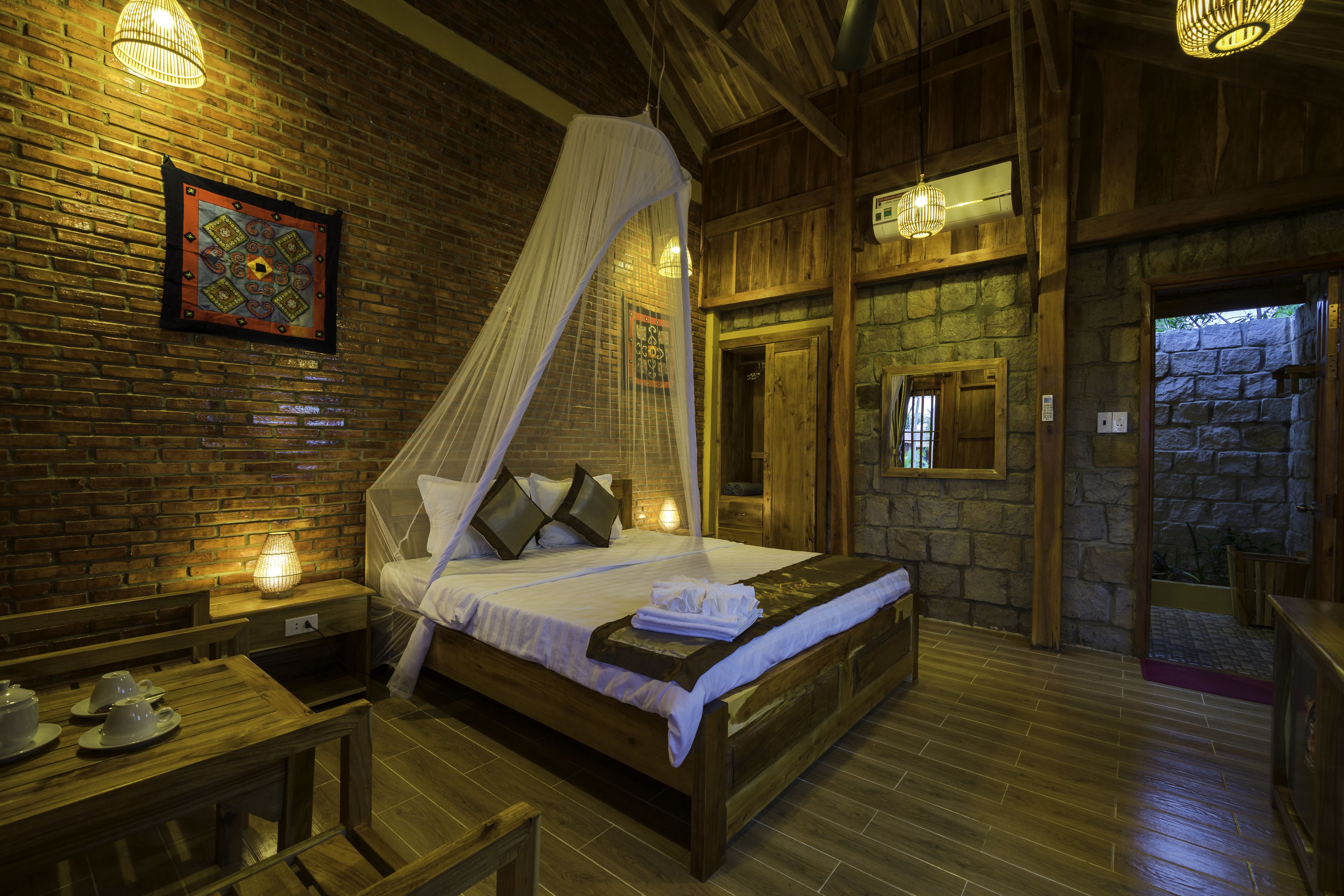 A wooden four-poster bed with a canopy, a bedside table with a lamp, and a brick wall with a decorative panel.