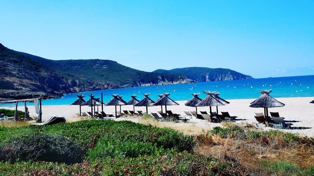 A beach with rows of sun loungers and thatched umbrellas, surrounded by hills and a clear blue sea.