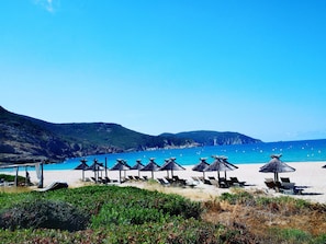 Ein Strand mit Liegestuhlreihen und Reetdachschirmen, umgeben von Hügeln und einem klaren, blauen Meer.