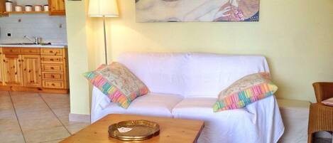Una sala de estar con un sofá blanco, almohadones de colores, una mesa de centro de madera, un cuadro en la pared y una cocina al fondo.