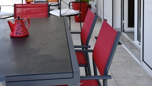 Un coin repas avec une longue table, des chaises rouges et une théière rouge. Il y a deux tableaux au mur et une plante en pot.