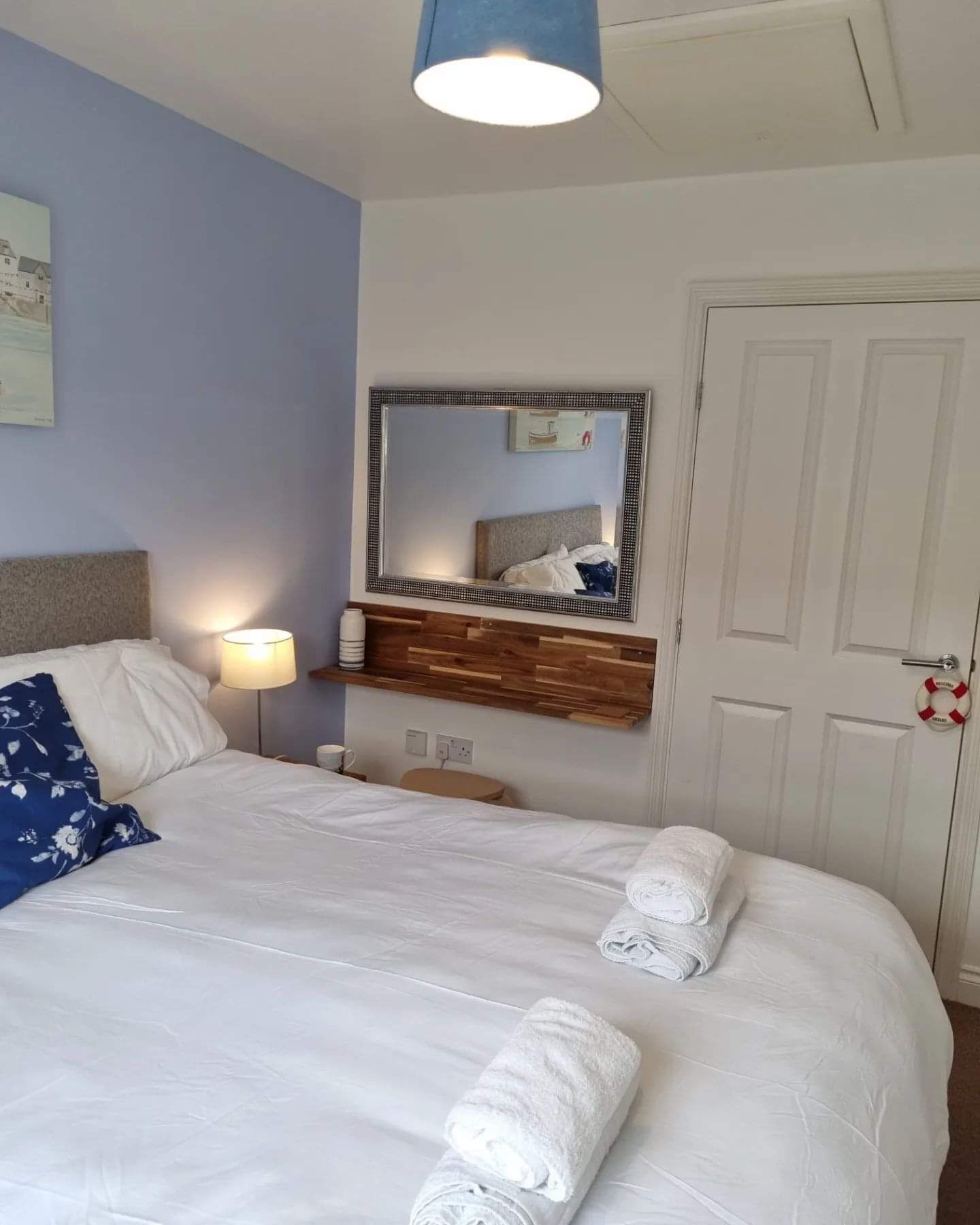 A neatly made bed with white linens and two rolled towels, a bedside lamp, a wooden headboard, a mirror with a decorative frame, and a white door with a life preserver hanging on it.
