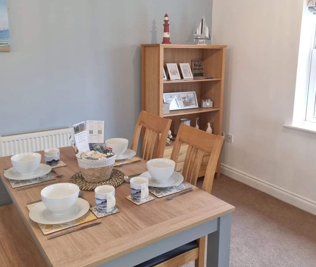 A dining area with a wooden table set for four, surrounded by chairs. A bookshelf with decorative items is visible in the background.