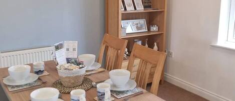 A dining area with a wooden table set for four, surrounded by chairs. A bookshelf with decorative items is visible in the background.
